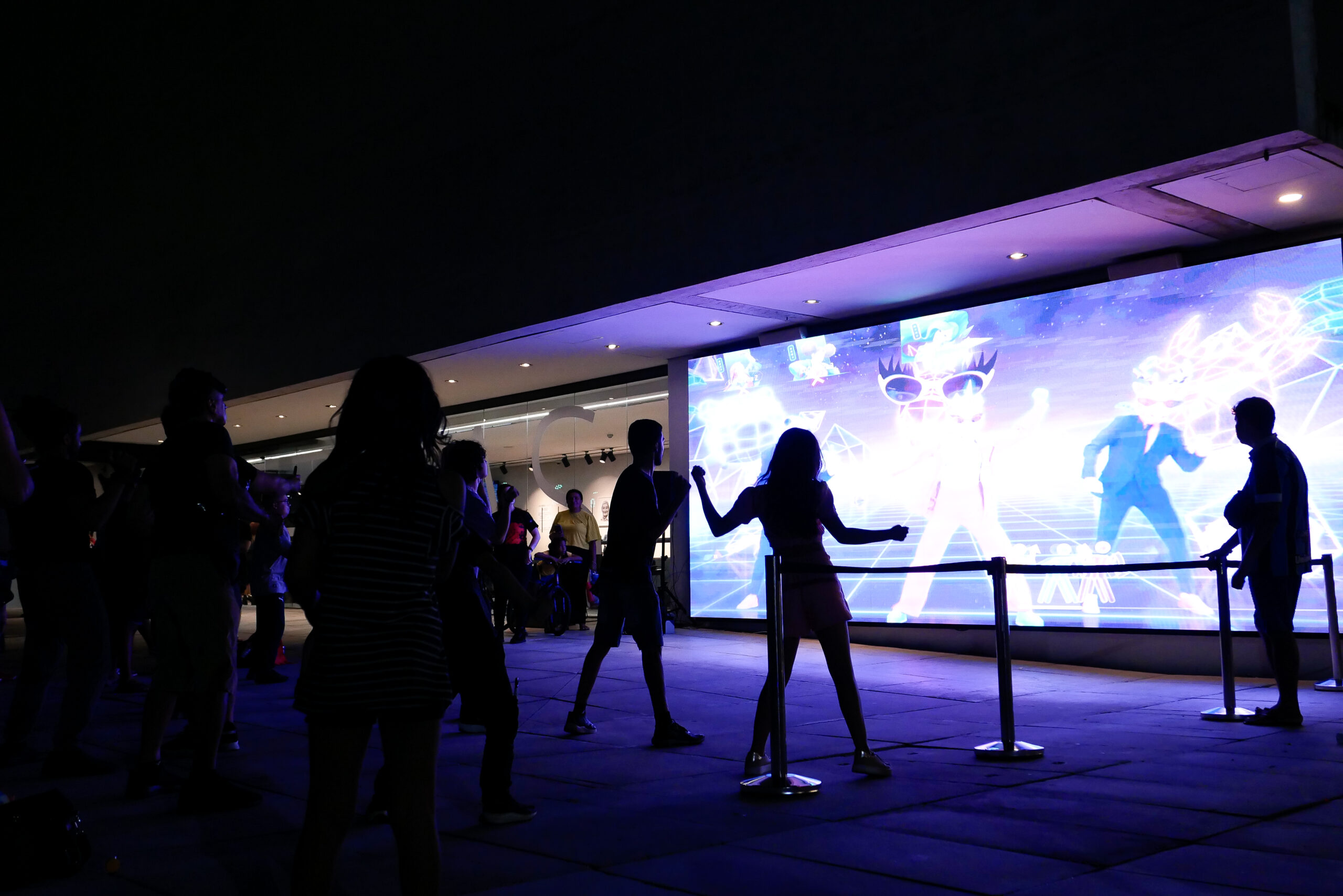 A imagem mostra pessoas interagindo em frente a uma grande tela iluminada em um ambiente externo à noite. A tela exibe gráficos coloridos e vibrantes, possivelmente de um jogo ou instalação interativa. As silhuetas das pessoas aparecem em destaque contra o brilho da projeção, sugerindo que estão participando de uma experiência digital ou tecnológica, como um jogo de realidade aumentada ou performance audiovisual interativa.