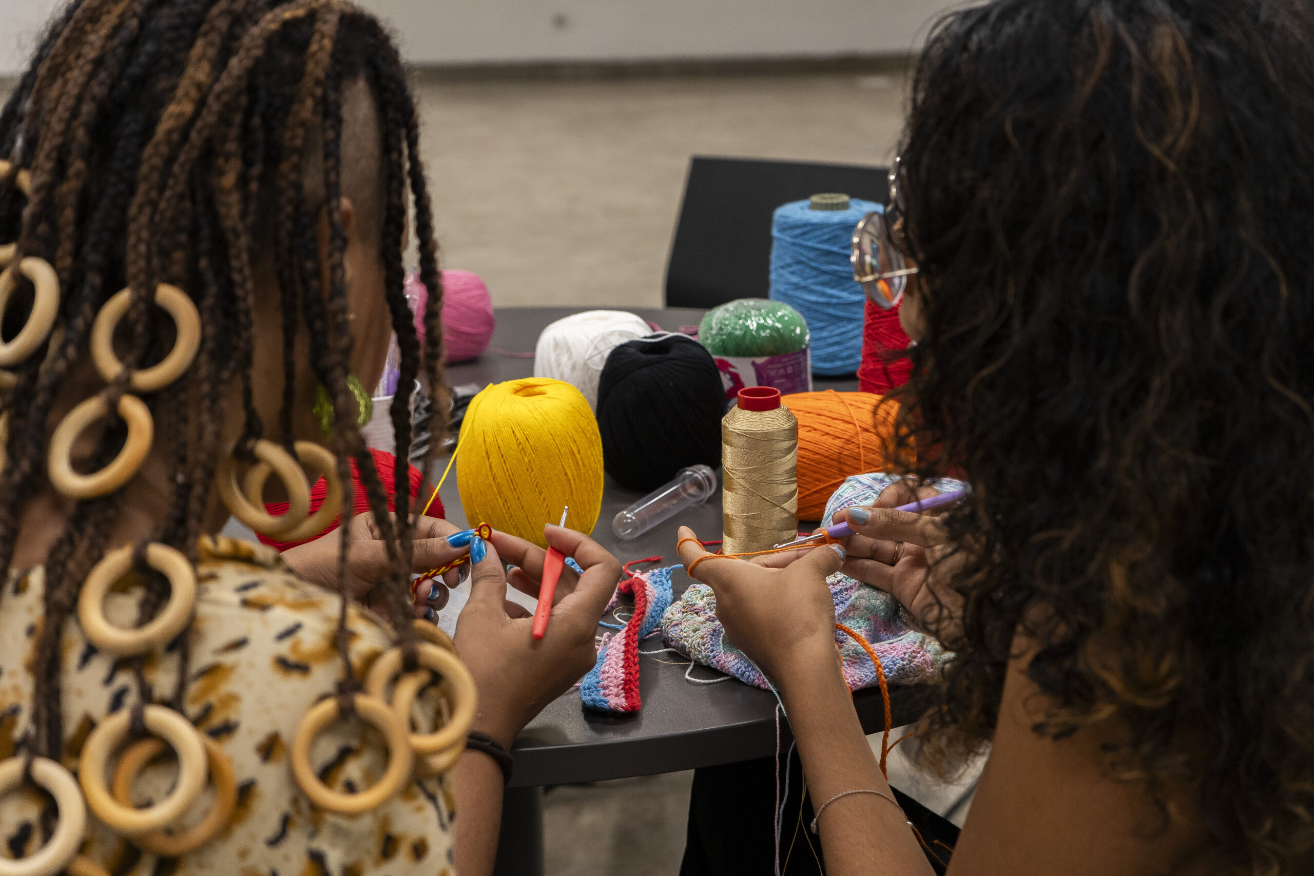 Duas mulheres estão sentadas lado a lado participando de uma atividade manual com fios, provavelmente crochê ou tricô. Elas estão de costas para a câmera, concentradas no trabalho com as mãos. Sobre a mesa à frente delas, há vários novelos de linhas coloridas (amarelo, branco, verde, rosa, vermelho, azul). A mulher à esquerda tem cabelos com tranças e adornos circulares de madeira, enquanto a da direita tem cabelos cacheados. O ambiente parece ser uma sala de oficina ou espaço cultural.