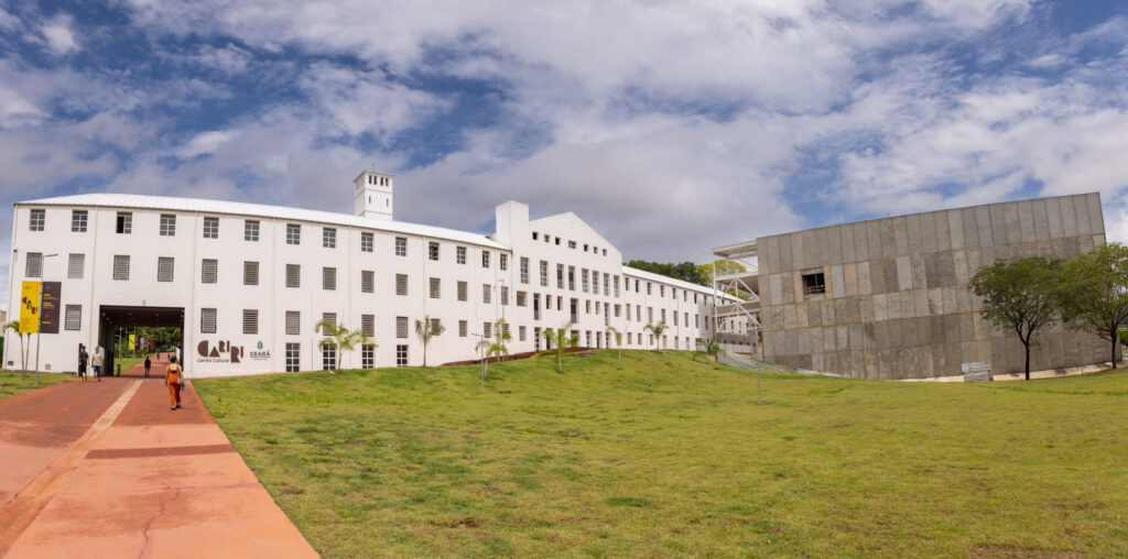 A imagem mostra uma construção ampla, com dois blocos contrastantes. À esquerda, um edifício branco de dois andares com janelas retangulares e arquitetura colonial moderna; à direita, uma estrutura contemporânea com fachada cinza e linhas retas. Em frente aos prédios, há um amplo gramado verde e uma calçada em tom terroso que conduz até a entrada do edifício branco. O céu está parcialmente nublado, com nuvens esparsas em meio ao azul. A imagem aparenta retratar um centro cultural, museu ou instituição pública moderna inserida em um espaço arquitetônico planejado.