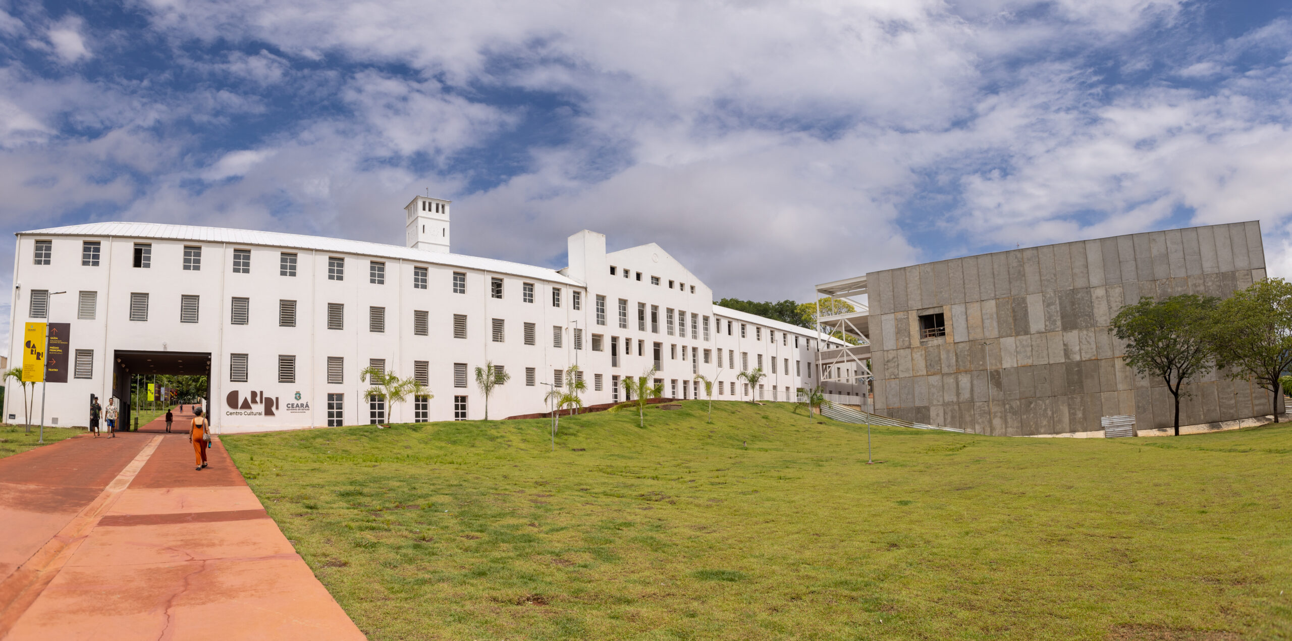 A imagem mostra uma construção ampla, com dois blocos contrastantes. À esquerda, um edifício branco de dois andares com janelas retangulares e arquitetura colonial moderna; à direita, uma estrutura contemporânea com fachada cinza e linhas retas. Em frente aos prédios, há um amplo gramado verde e uma calçada em tom terroso que conduz até a entrada do edifício branco. O céu está parcialmente nublado, com nuvens esparsas em meio ao azul. A imagem aparenta retratar um centro cultural, museu ou instituição pública moderna inserida em um espaço arquitetônico planejado.